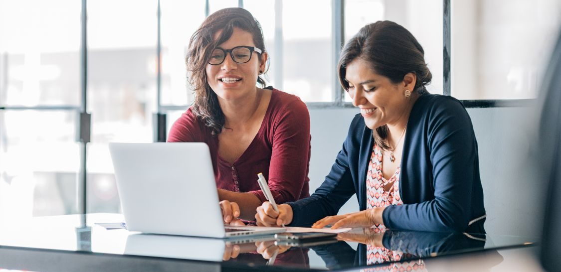 two women sit at a desk, one looking at a laptop while the other writes something