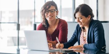 two women sit at a desk, one looking at a laptop while the other writes something