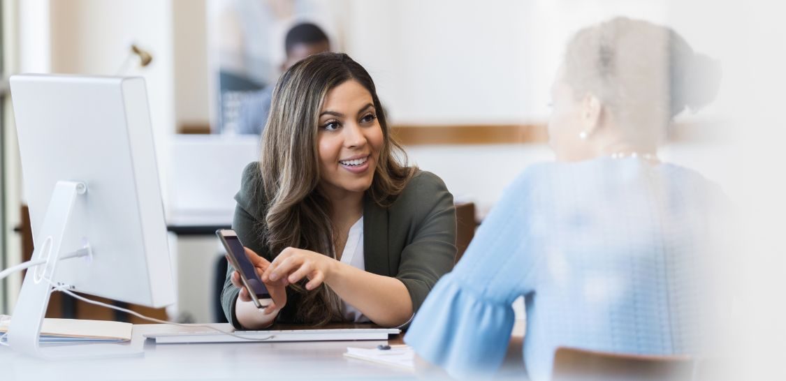 A woman with mid-length brown hair wearing a suit jacket sits behind a desk that has a computer monitor on it. She is showing her cell phone screen to a woman sitting on the near side of the desk.