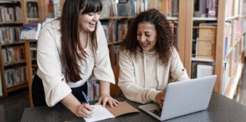 Two women in a library at a desk, one standing and writing on a notepad while the other sits at a laptop. They are both smiling.