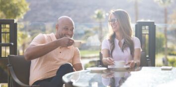 A man and a woman sit at an outside table drinking from teacups with a beautiful landscape in the background