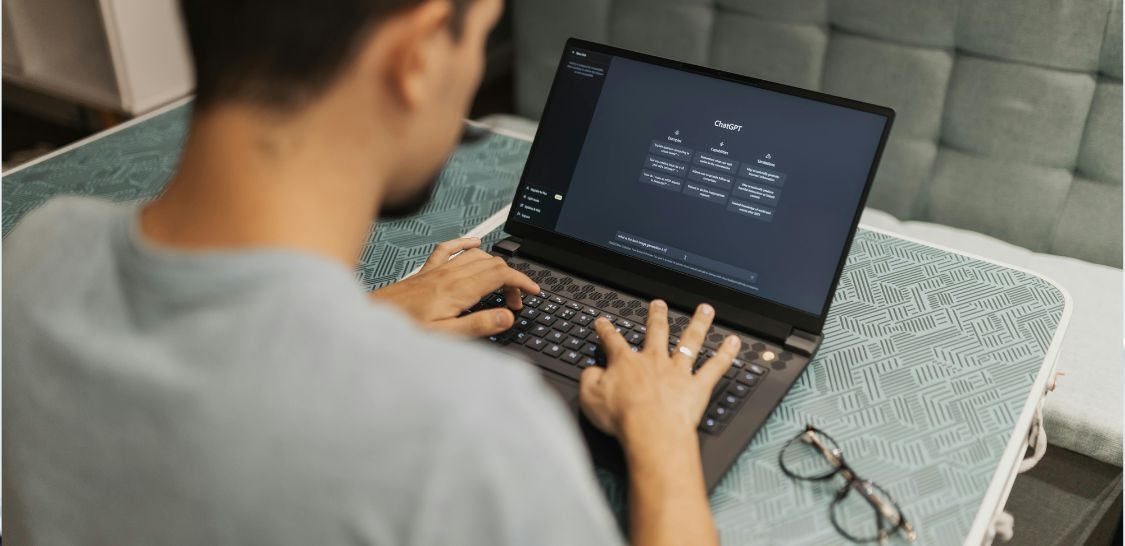 Photographed from over the shoulder, a man sits at a table typing on a laptop keyboard with his eye glasses sitting to the right of the laptop. The screen shows Chat GPT.