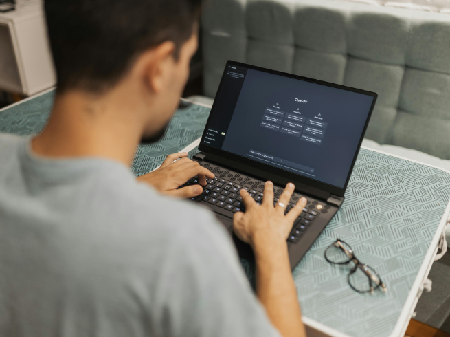 Photographed from over the shoulder, a man sits at a table typing on a laptop keyboard with his eye glasses sitting to the right of the laptop. The screen shows Chat GPT.