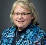 Headshot of Denise Hale. A woman with glasses, shoulder length blonde hair with bangs. She is wearing a blouse with various shades of blue, floral pattern, and a navy blue shirt underneath.