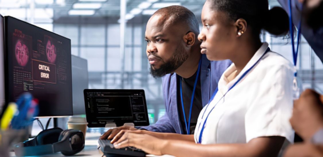 And man and a woman sit at a desk in front of a computer screen that has the words "Critical Breach" on it.