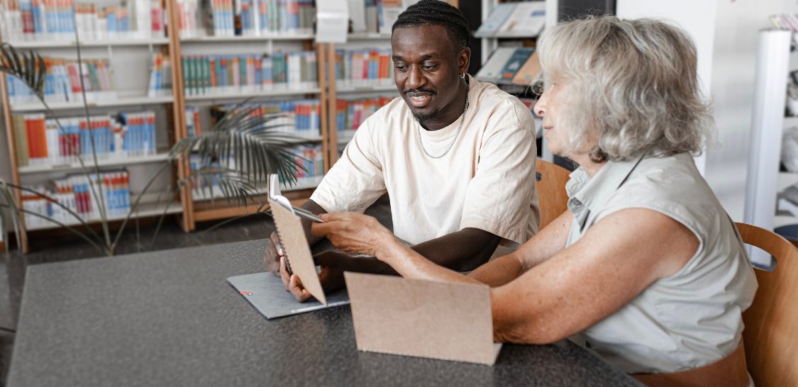 A man and woman sit at a table, both looking at tablets, but they are clearly collaborating. Shelves full of books sit in the background.