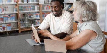 A man and woman sit at a table, both looking at tablets, but they are clearly collaborating. Shelves full of books sit in the background.