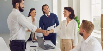 A group of people stand around a table and a man and a woman are shaking hands across the table.
