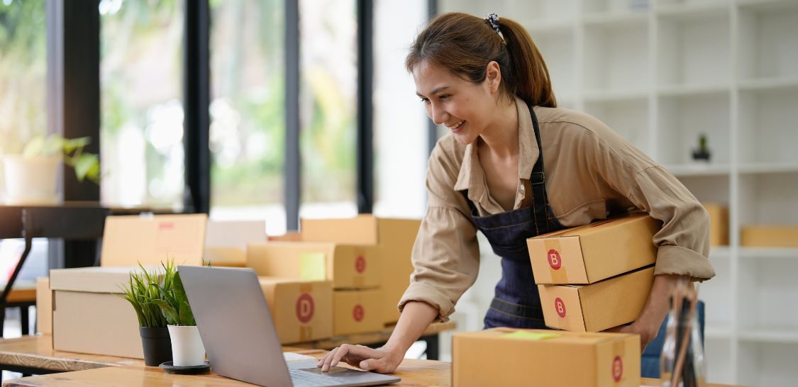 A woman types on a laptop while standing at a table with boxes behind her and to her right. She is holding two boxes under her right arm. She is shipping items.
