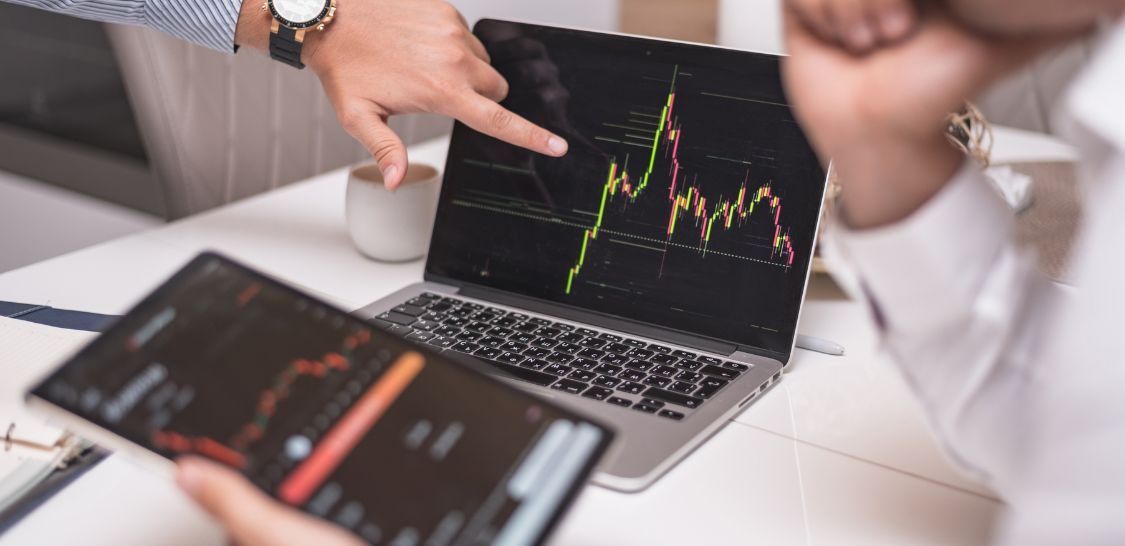 A hand points at a laptop screen that shows a graph of the stock market and a person in the right side of the image holds a tablet with similar information on the screen and his chin in his hand like he's thinking