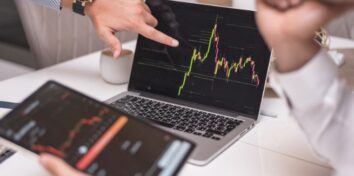 A hand points at a laptop screen that shows a graph of the stock market and a person in the right side of the image holds a tablet with similar information on the screen and his chin in his hand like he's thinking