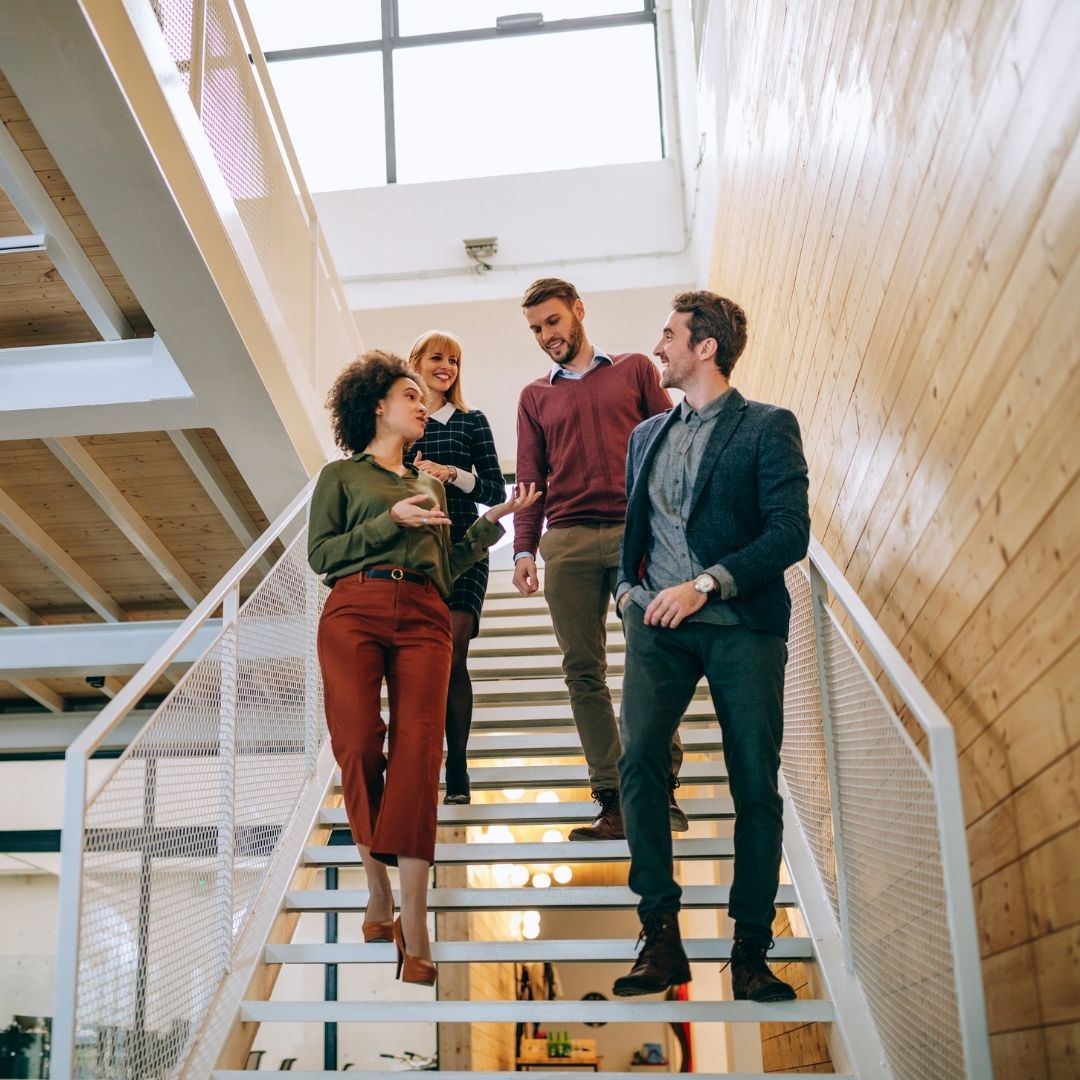 Four colleagues discuss business operations while walking down a staircase in a modern office.