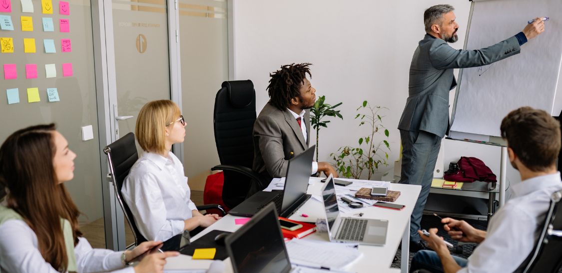 Five colleagues attend a meeting as a man presents at a whiteboard, with laptops, documents, and sticky notes spread across the conference table.