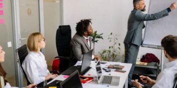 Five colleagues attend a meeting as a man presents at a whiteboard, with laptops, documents, and sticky notes spread across the conference table.