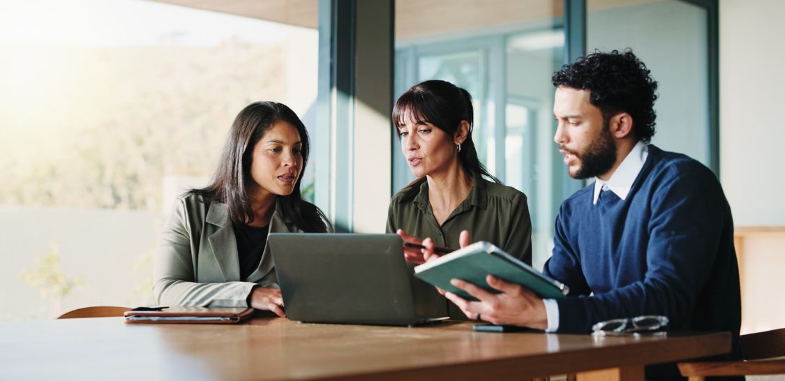 Three colleagues review information on a laptop and tablet during a meeting in a modern office.