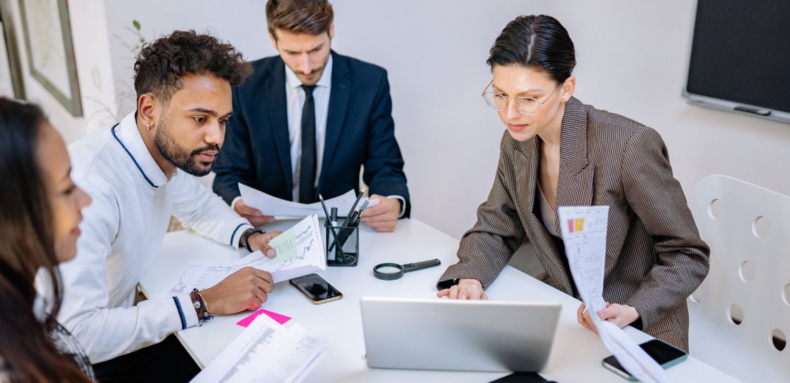 A group of people sit around a desk with papers scattered around. They are looking at a laptop.