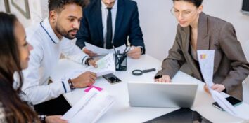 A group of people sit around a desk with papers scattered around. They are looking at a laptop.