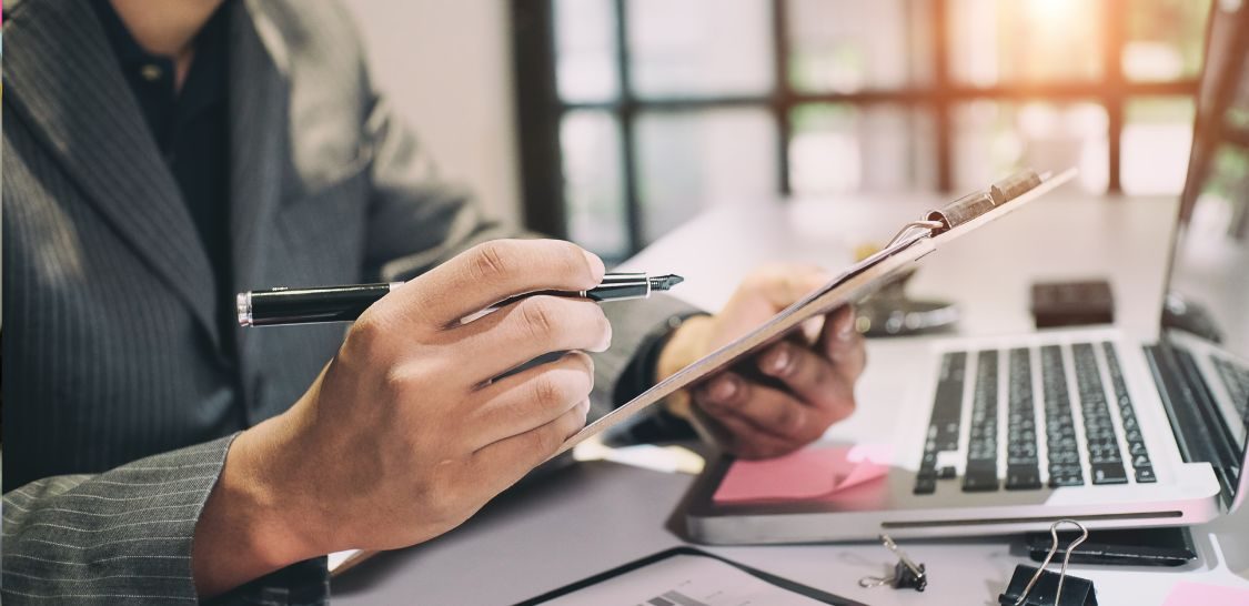 Person reviewing documents on a clipboard beside a laptop, financial charts, and office supplies.