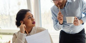 Two professionals discuss a document while reviewing workplace information in an office.