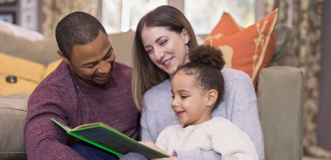 Parents and young child read a book together at home.