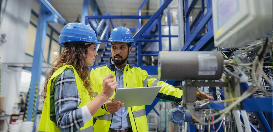 Two manufacturing professionals wearing hard hats and high-visibility jackets review a tablet beside industrial equipment on a factory floor.