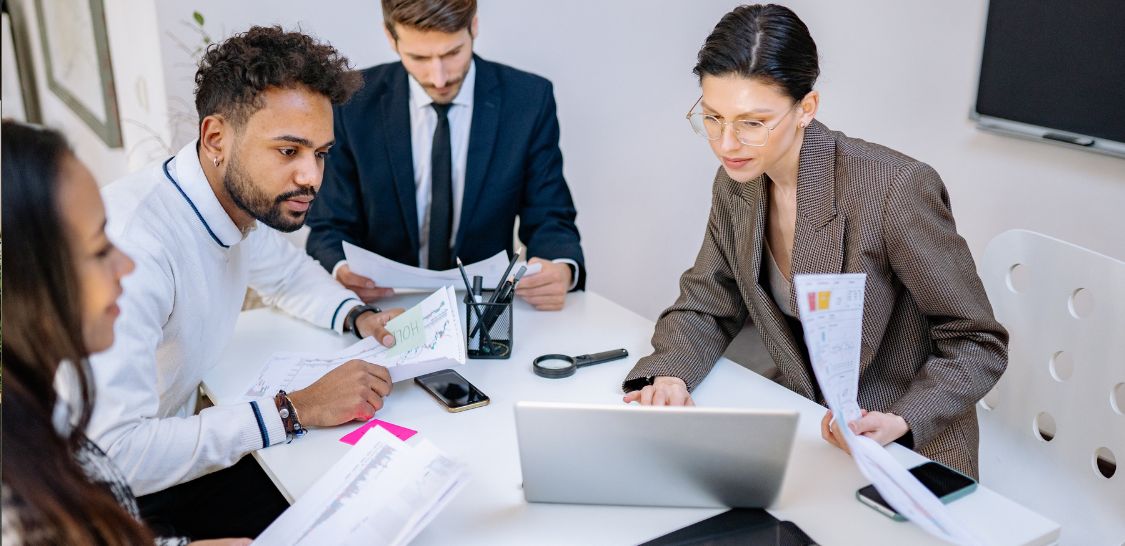 a group of professional sit around a table looking at a laptop and papers