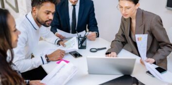 a group of professional sit around a table looking at a laptop and papers
