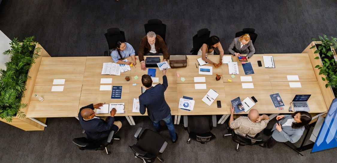 Overhead view of a diverse team meeting around a conference table, reviewing financial documents, charts, and laptops.