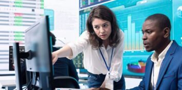 Two financial professionals review market data on computer screens in a trading office.